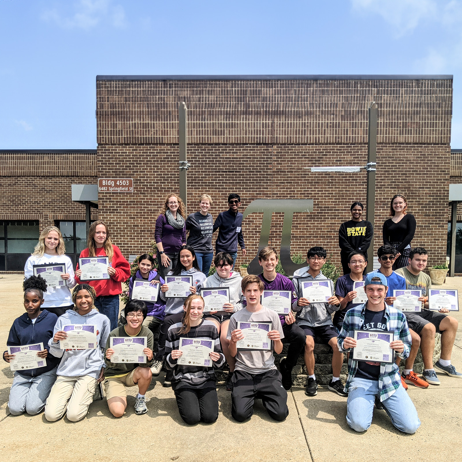 Group of students posing with GEMS AEOP certificates of completion, teachers in the background in front of a school building.
