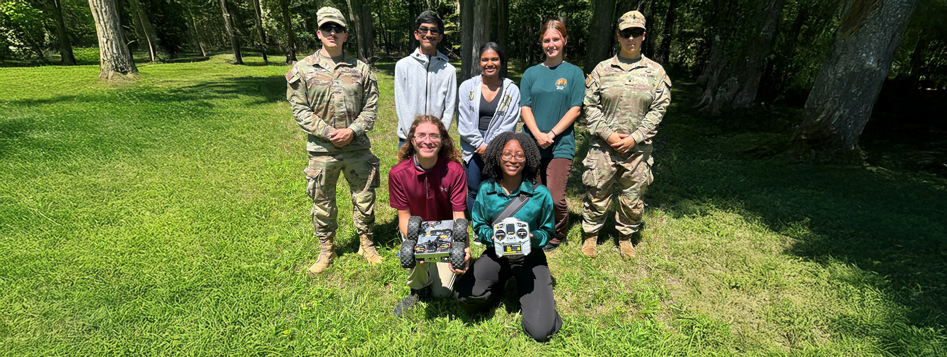 Photo of student interns holding a four-wheeled robot and a remote controller, flanked by US Army Uniformed Personnel.