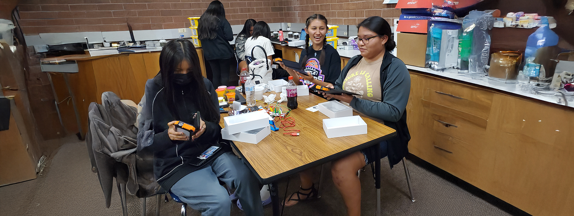 Two groups of students in a classroom open packages containing solar-powered battery banks which will allow them to charge their devices.